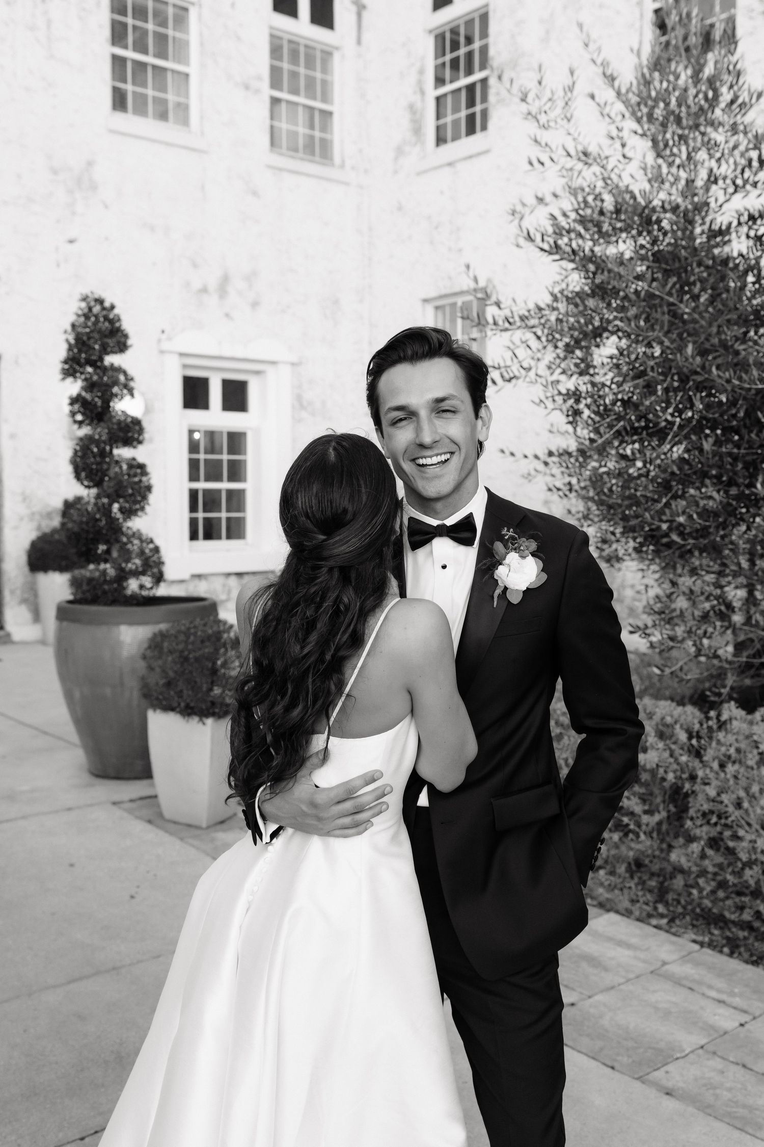 A black and white wedding photo showing a couple sharing a joyful moment outside a white building with elegant windows.
