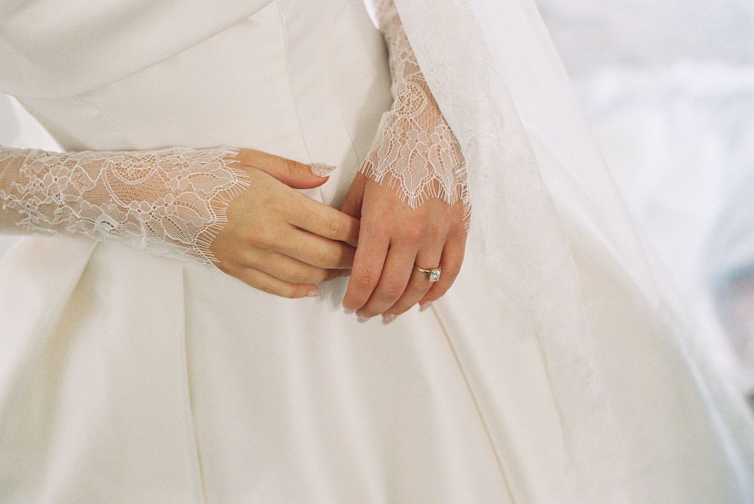 Bride in white lace wedding gown with elegant long sleeves, hands clasped together showing a diamond ring.