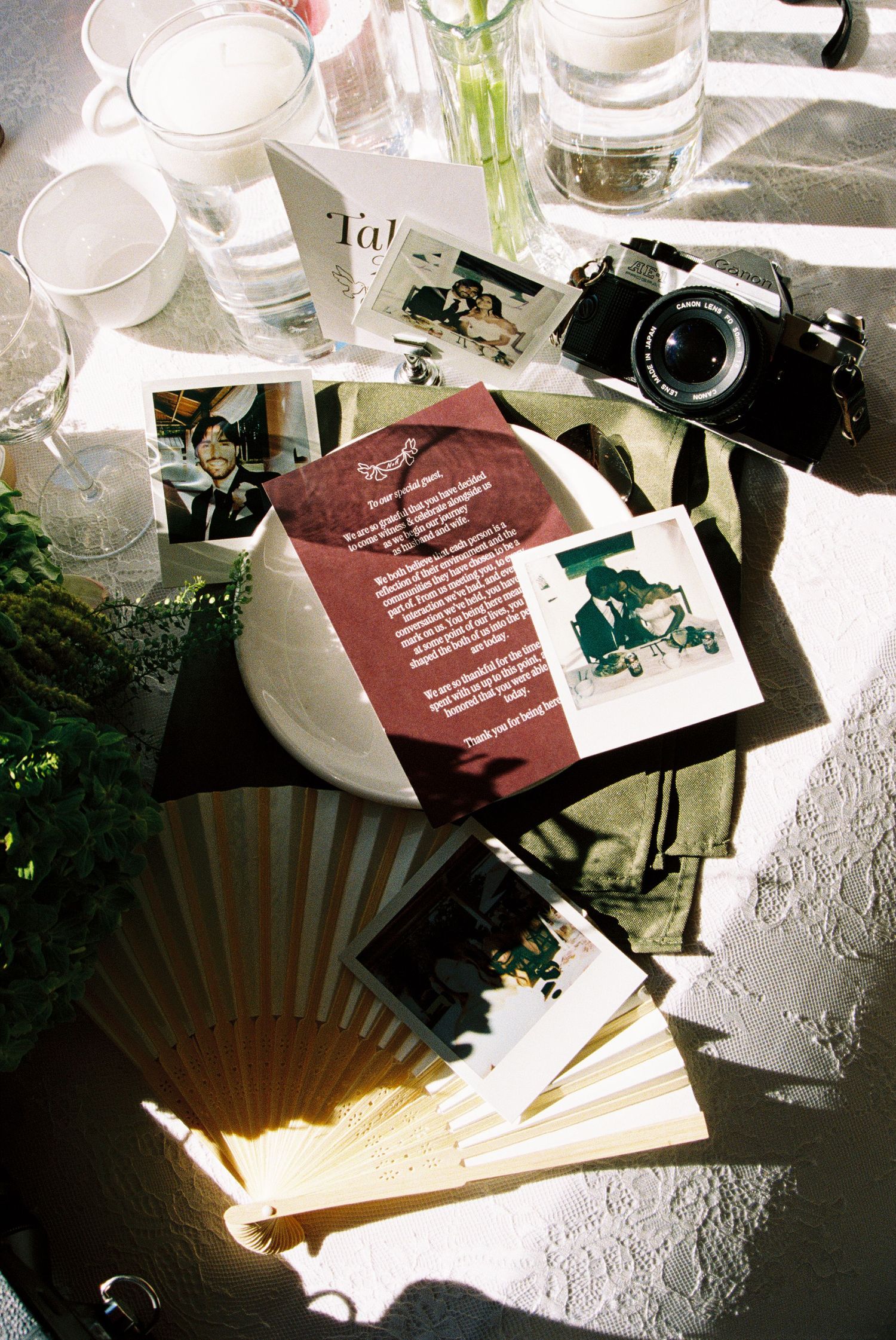 A vintage film camera, polaroid photos, a folding fan, and glassware arranged on a sunlit table at a wedding reception.