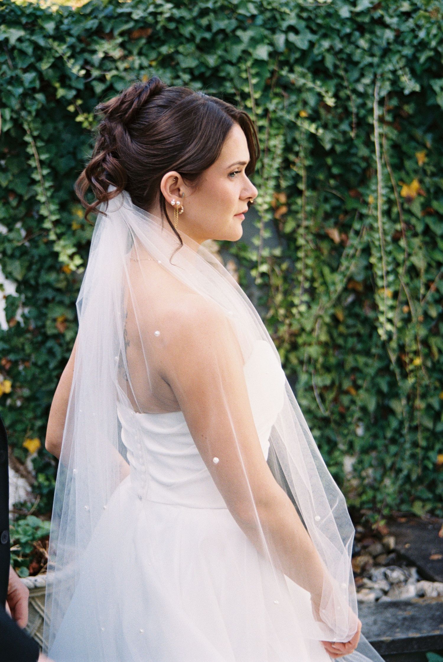 Bride in flowing white gown and veil against lush green ivy backdrop.