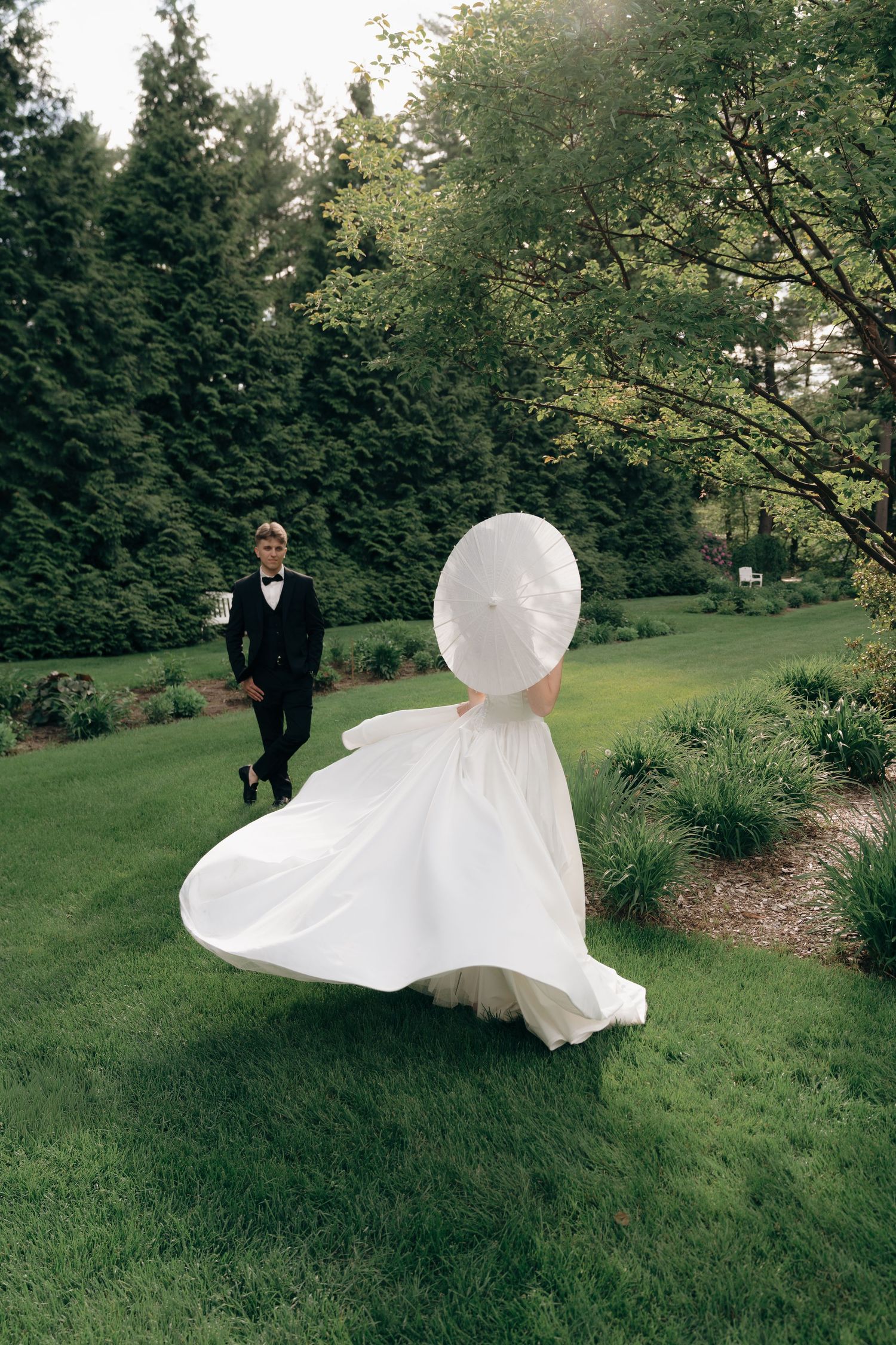 A flowing white wedding dress twirls gracefully on a manicured lawn surrounded by evergreen trees.