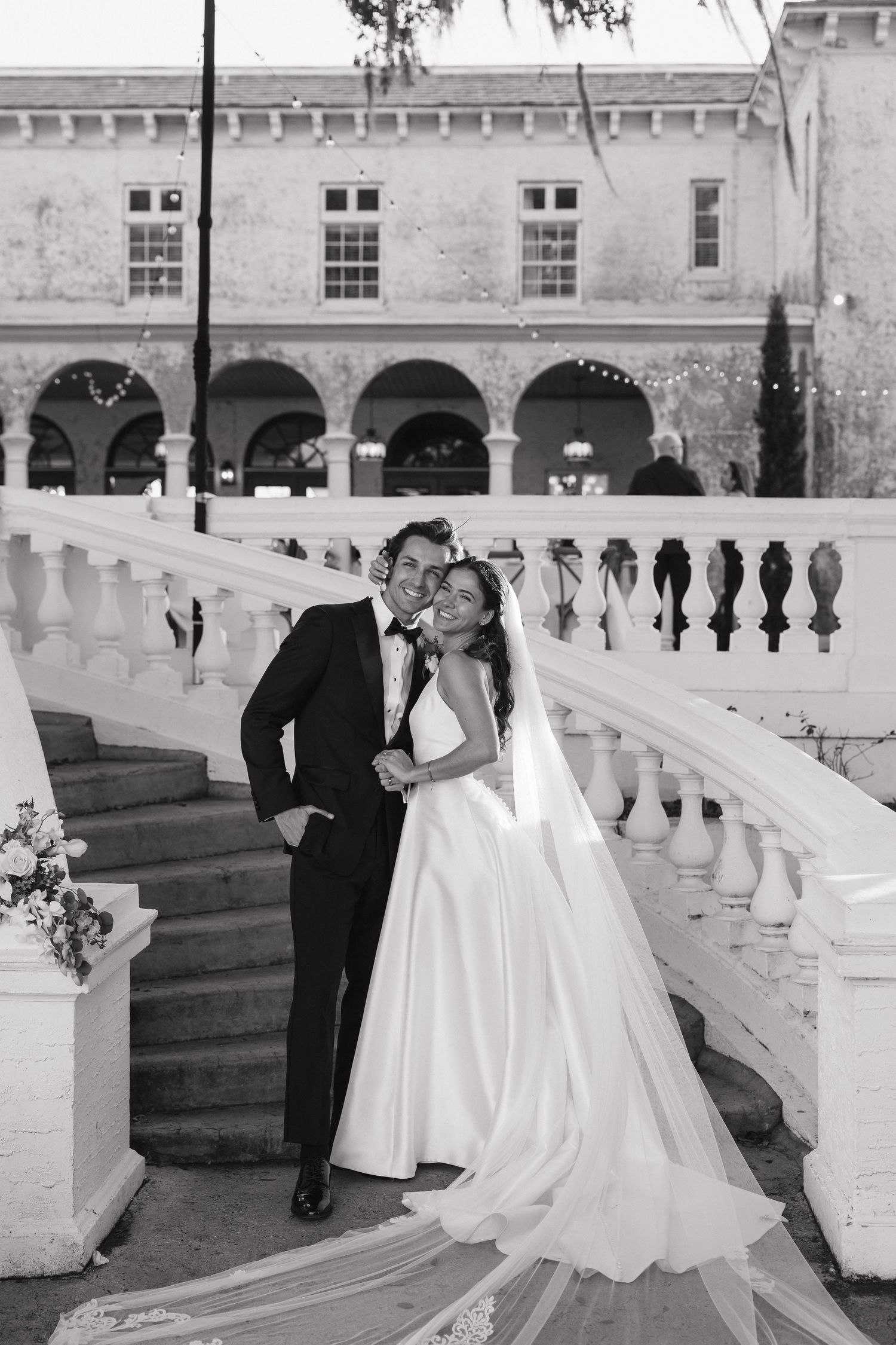 Wedding party in formal attire gathered on white stone balcony of historic building.