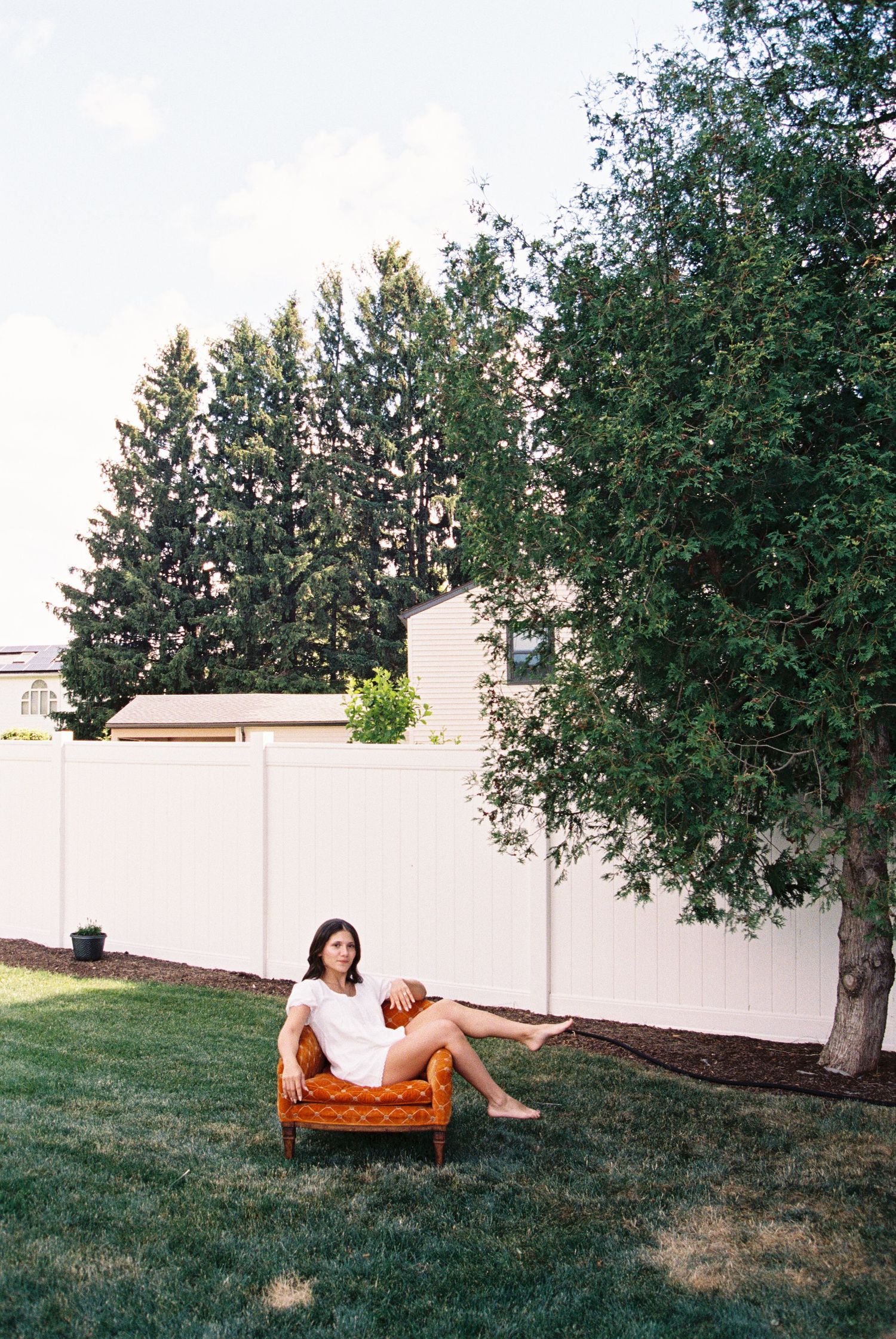 Woman in white dress relaxing on vintage orange chair in lush green backyard near tree and white fence.