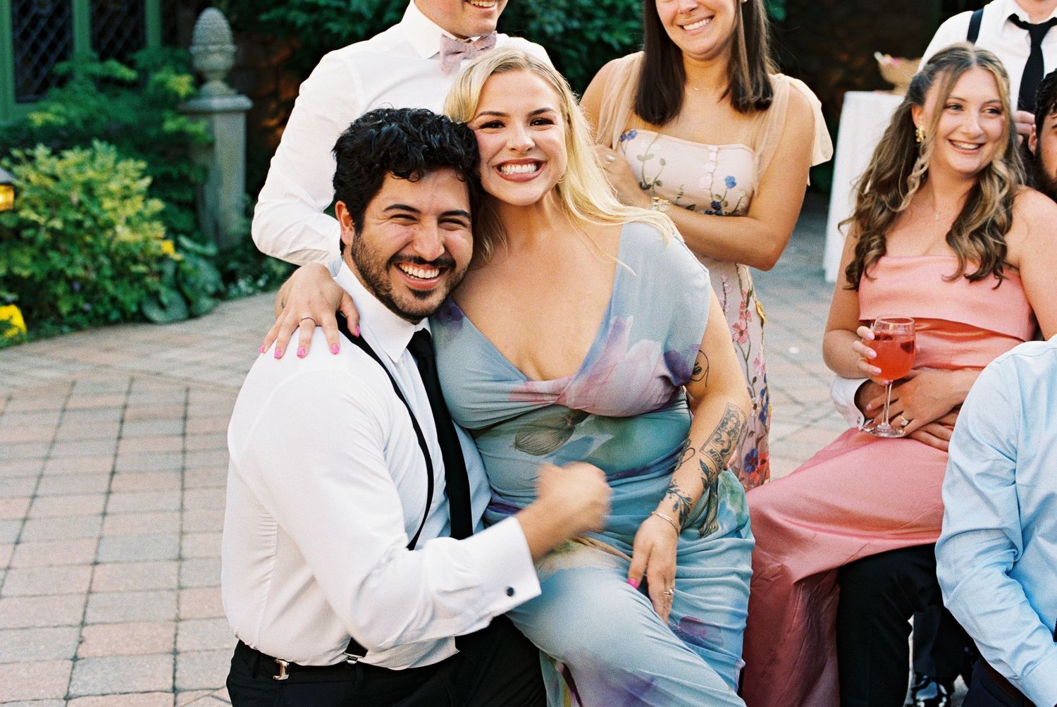Couple smiling and posing together at an outdoor wedding reception with guests in background.