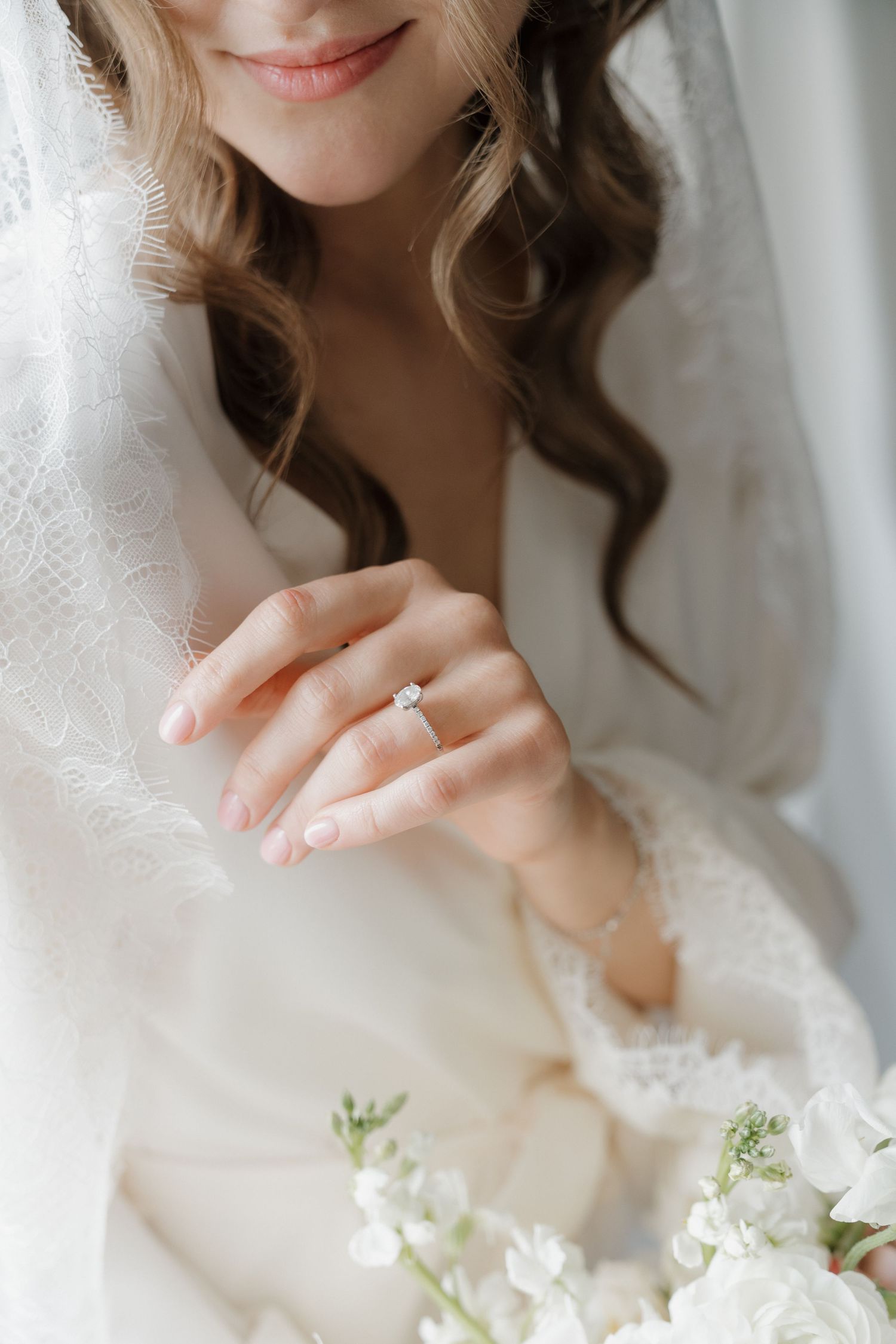 Close up of a delicate engagement ring on a hand with white wedding dress and bouquet details in soft natural light.