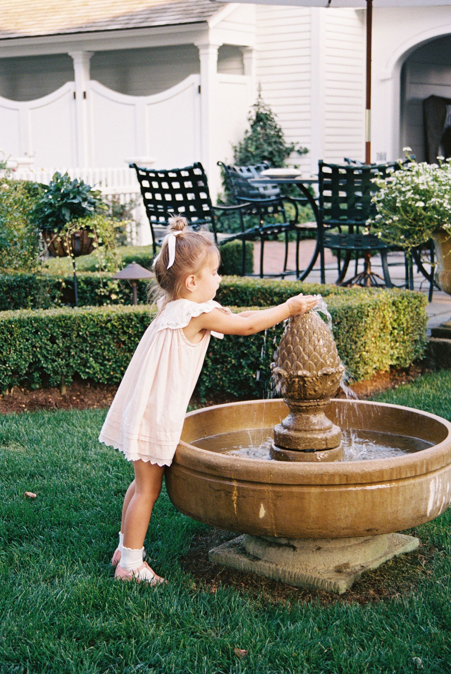 A small child in a white dress plays with a garden fountain on a manicured lawn with patio furniture in the background.