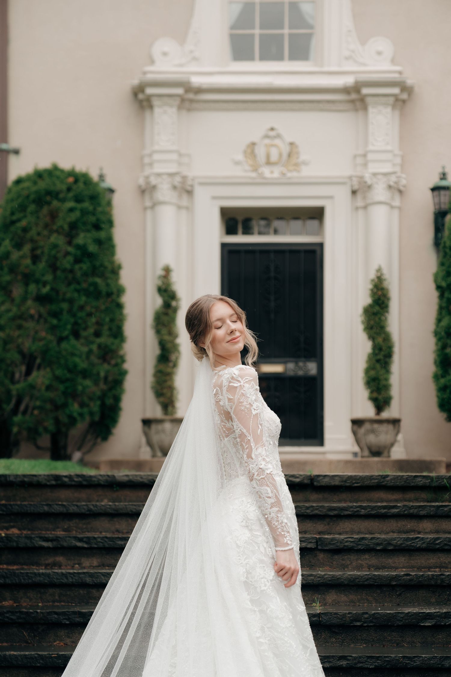 Elegant bride in lace long-sleeve wedding gown with flowing veil posing on steps outside classic mansion.