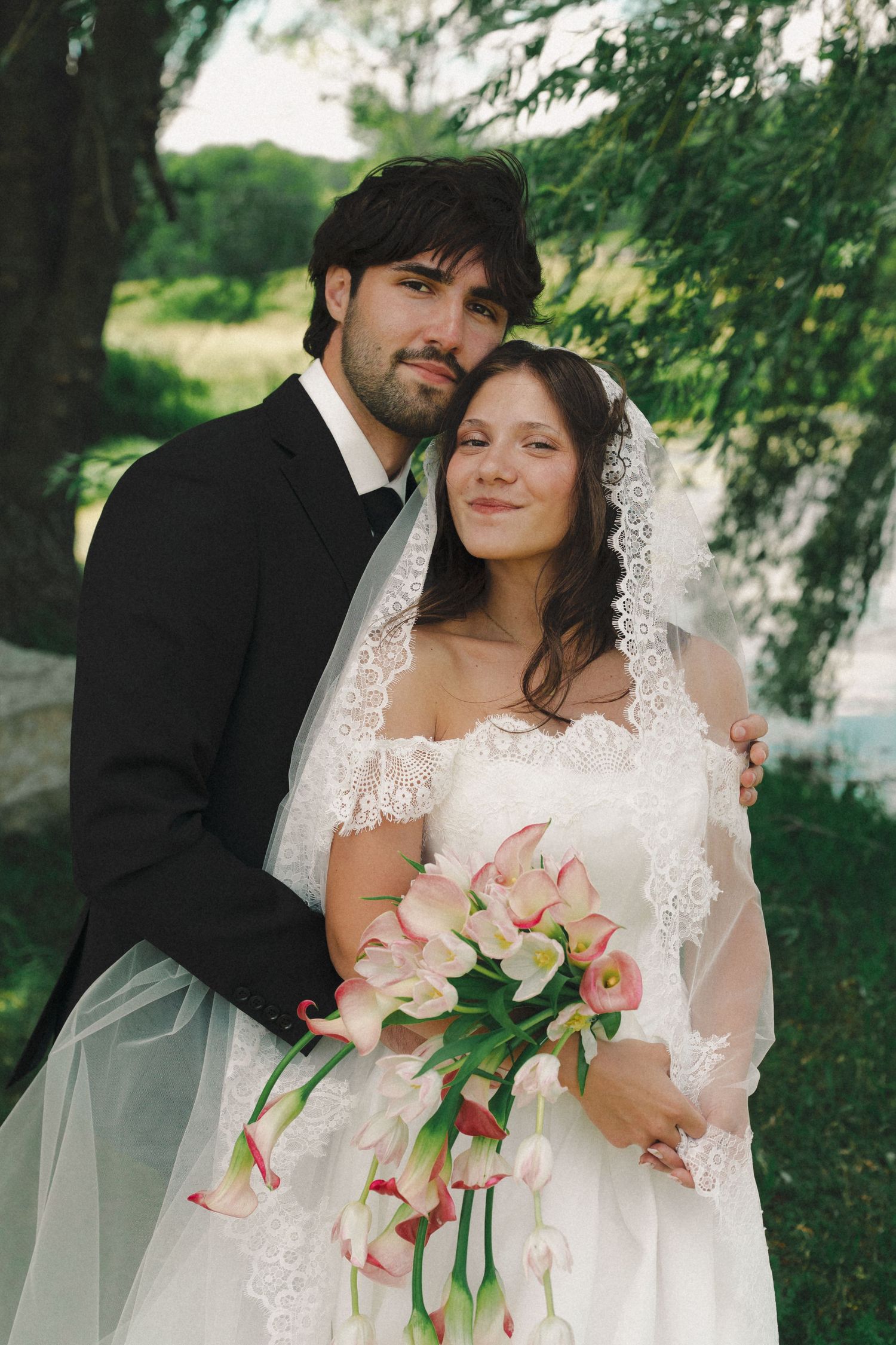 Bride and groom pose together outdoors, she holds pink calla lilies bouquet wearing lace veil and off-shoulder gown.