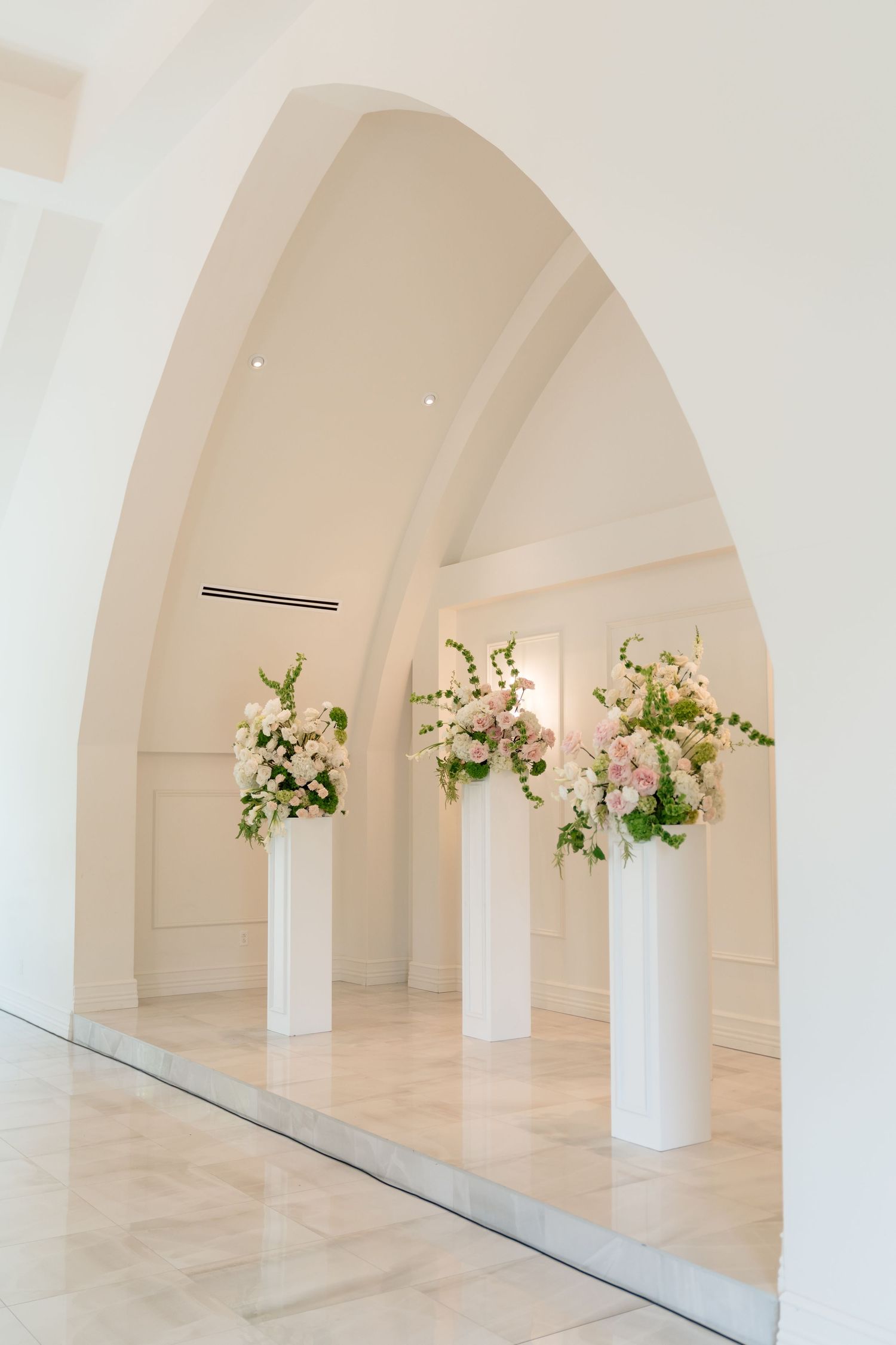 Three white pedestals with pink and white floral arrangements displayed under an elegant arched white interior alcove.