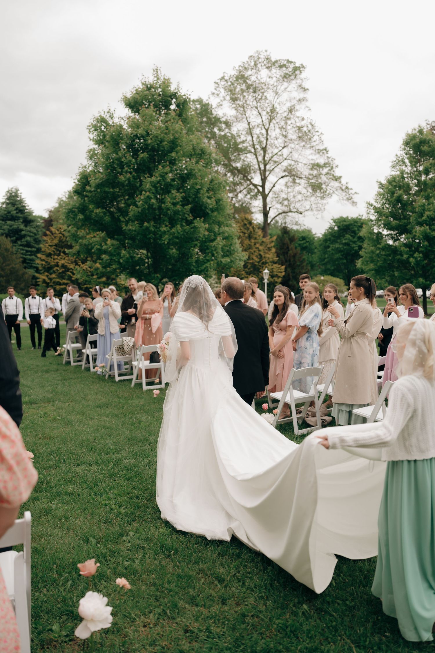 Wedding party members hold up the bride's long white dress train as they walk across a grassy outdoor ceremony venue.