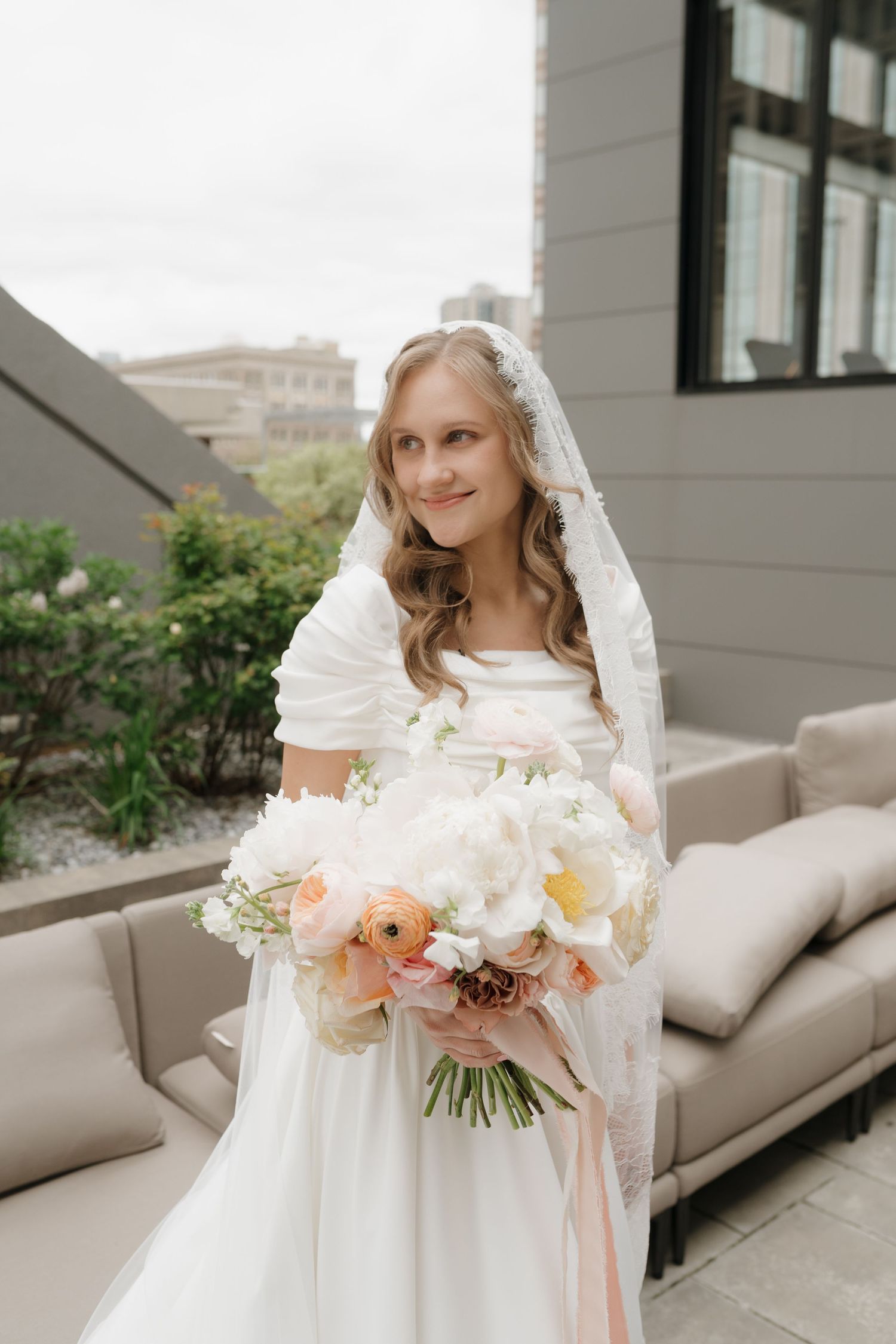 A bride in a white wedding dress holds a delicate bouquet of peach and white flowers with a flowing veil.