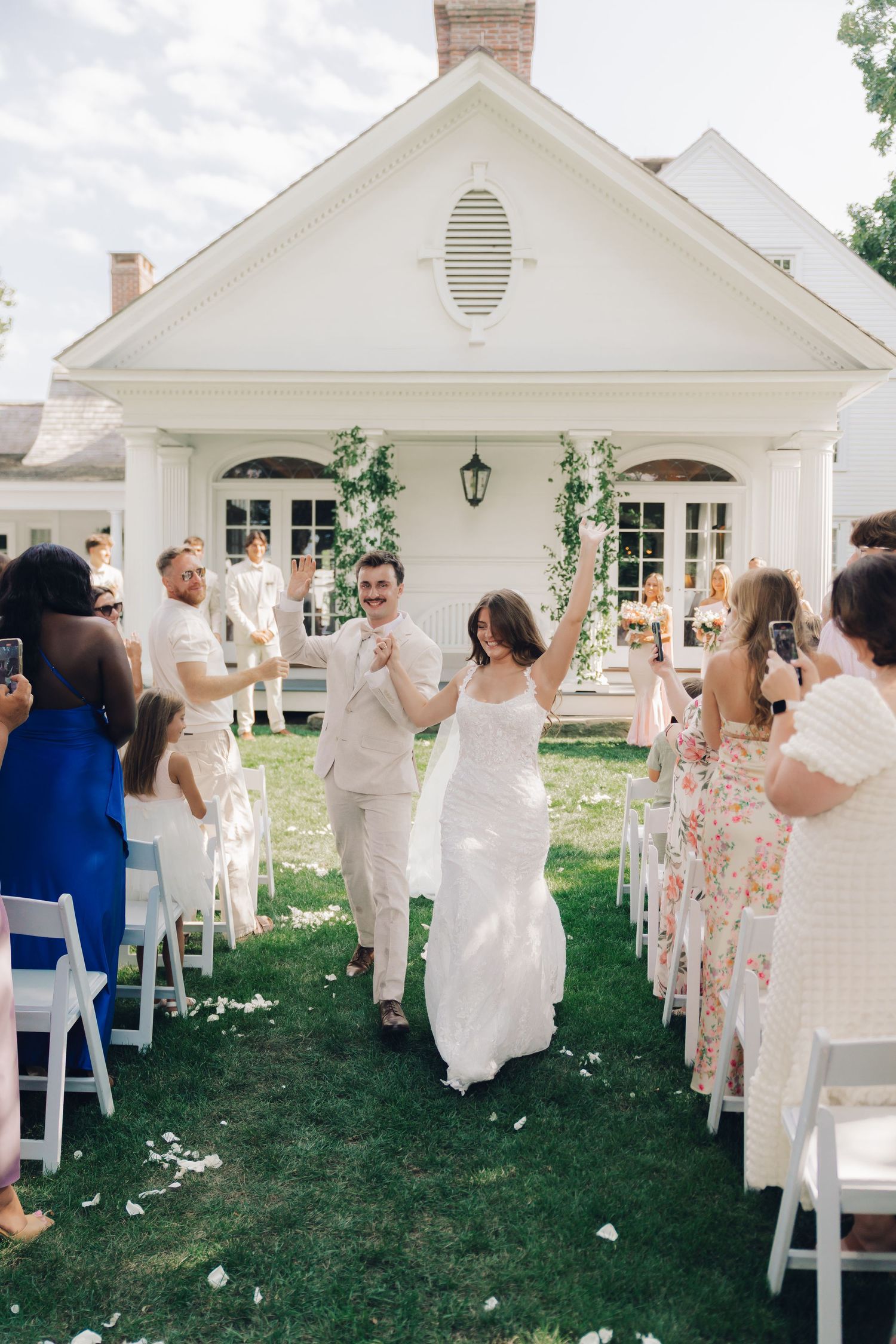 Wedding guests in white attire wave napkins during an outdoor celebration on a lawn with white chairs and buildings.