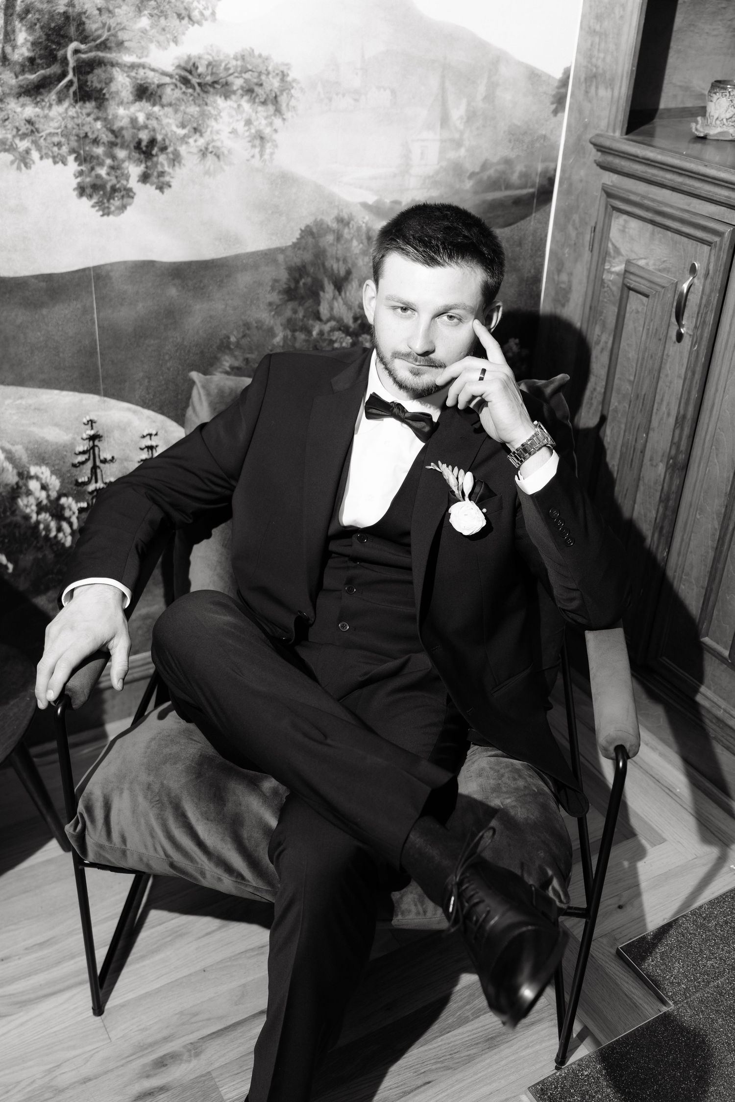 Black and white photo of a well-dressed groom in a tuxedo sitting in a chair, posing confidently with a boutonniere.