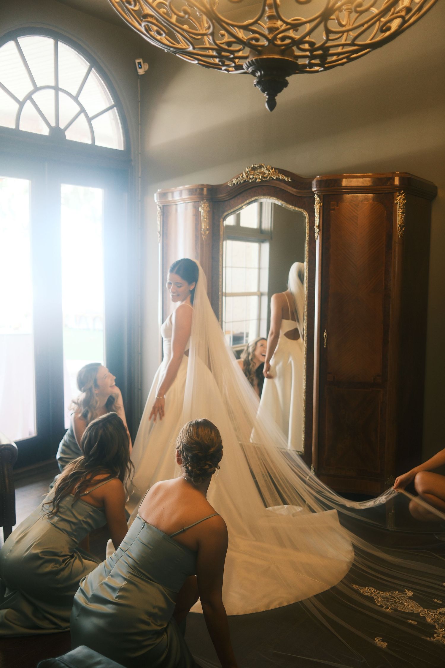 Wedding dress being prepared in an elegant room with ornate chandelier and mirrors.