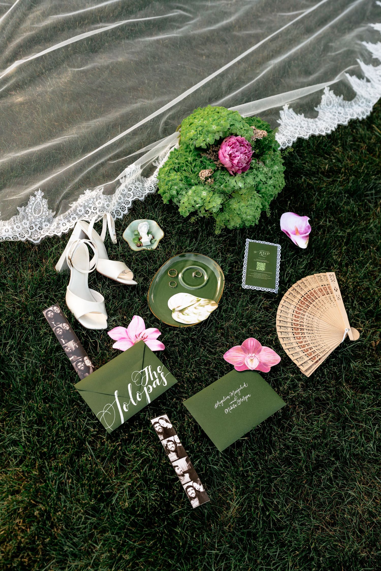 Wedding flat lay on grass with green stationery, white heels, veil, pink flowers, fan, and ring dish.