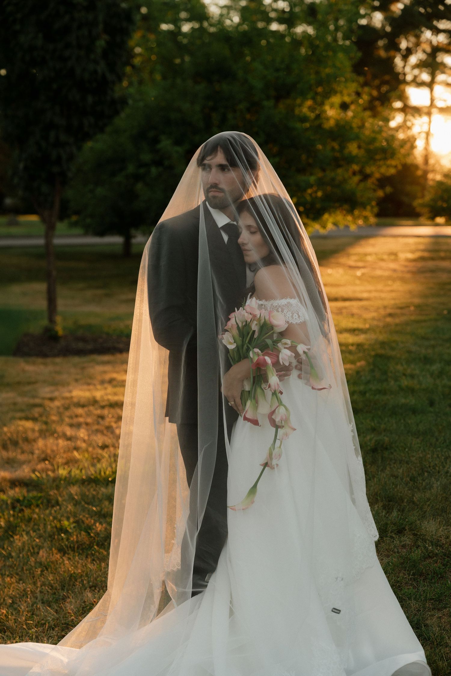 Bride and groom sharing intimate moment under flowing veil at golden sunset in lush green park.