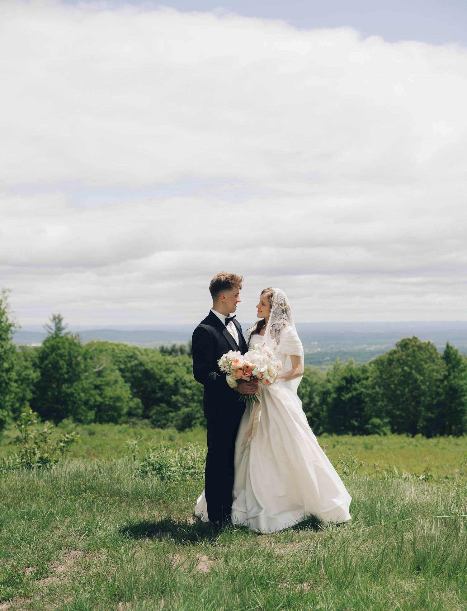 A couple in wedding attire stands together on a hilltop meadow with scenic mountain views in the background.