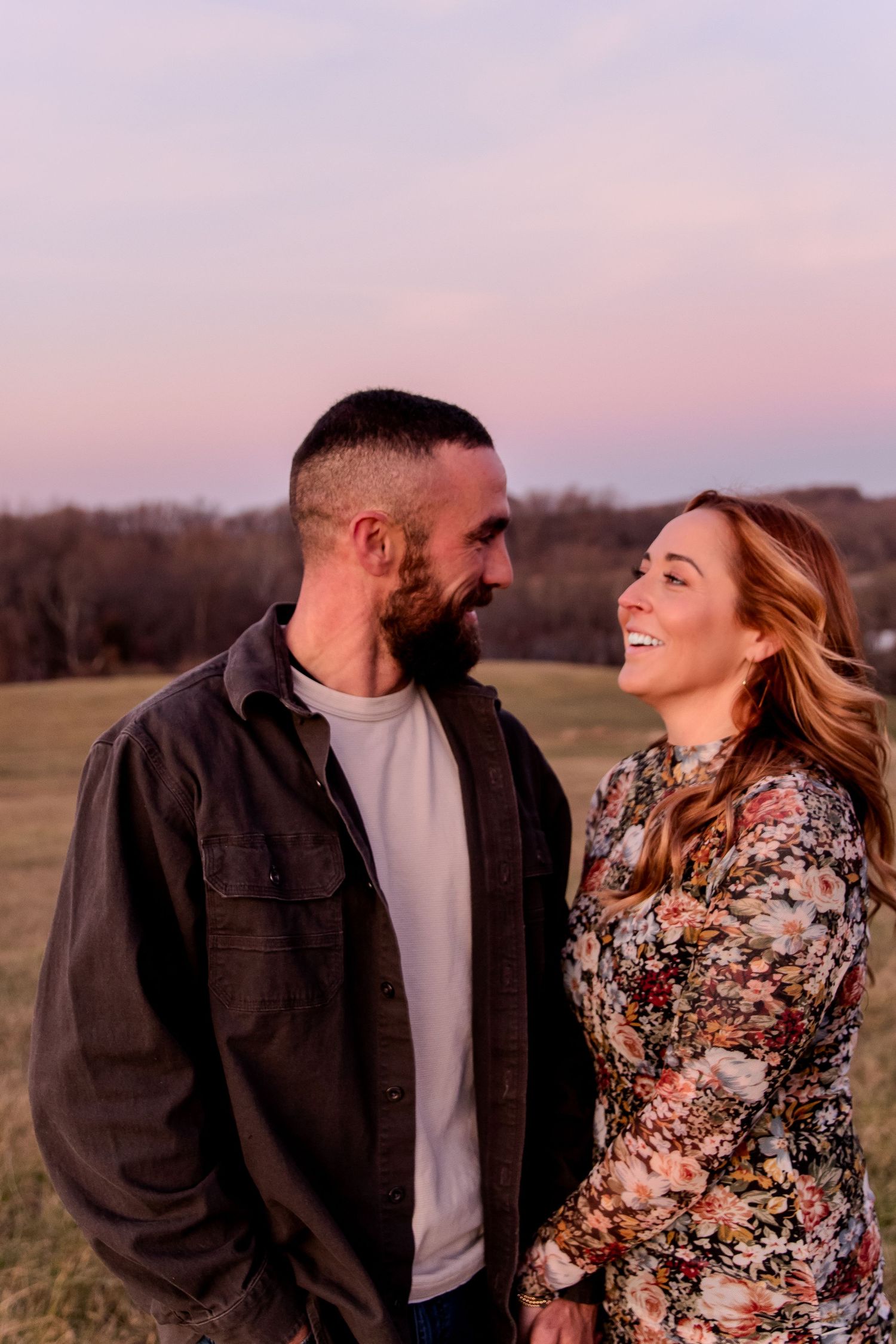 Couple smiling at each other during golden hour sunset in a green field, man in dark jacket, woman in floral dress.