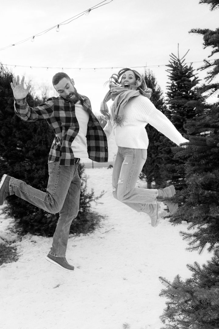 Joyful couple jumping in snowy Christmas tree farm, black and white winter photo.