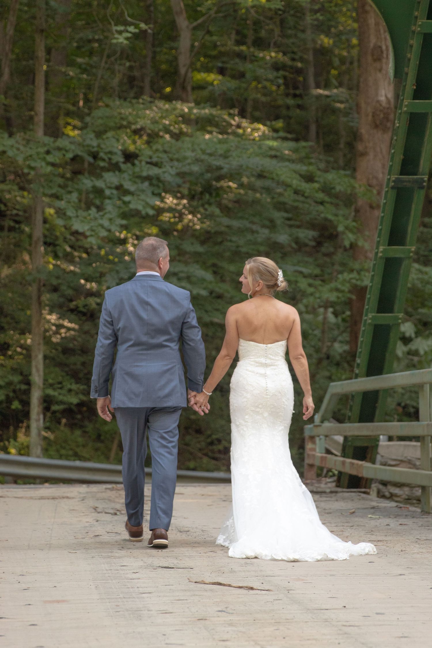Bride and groom walking away hand in hand on wooden bridge with forest scenery behind them.