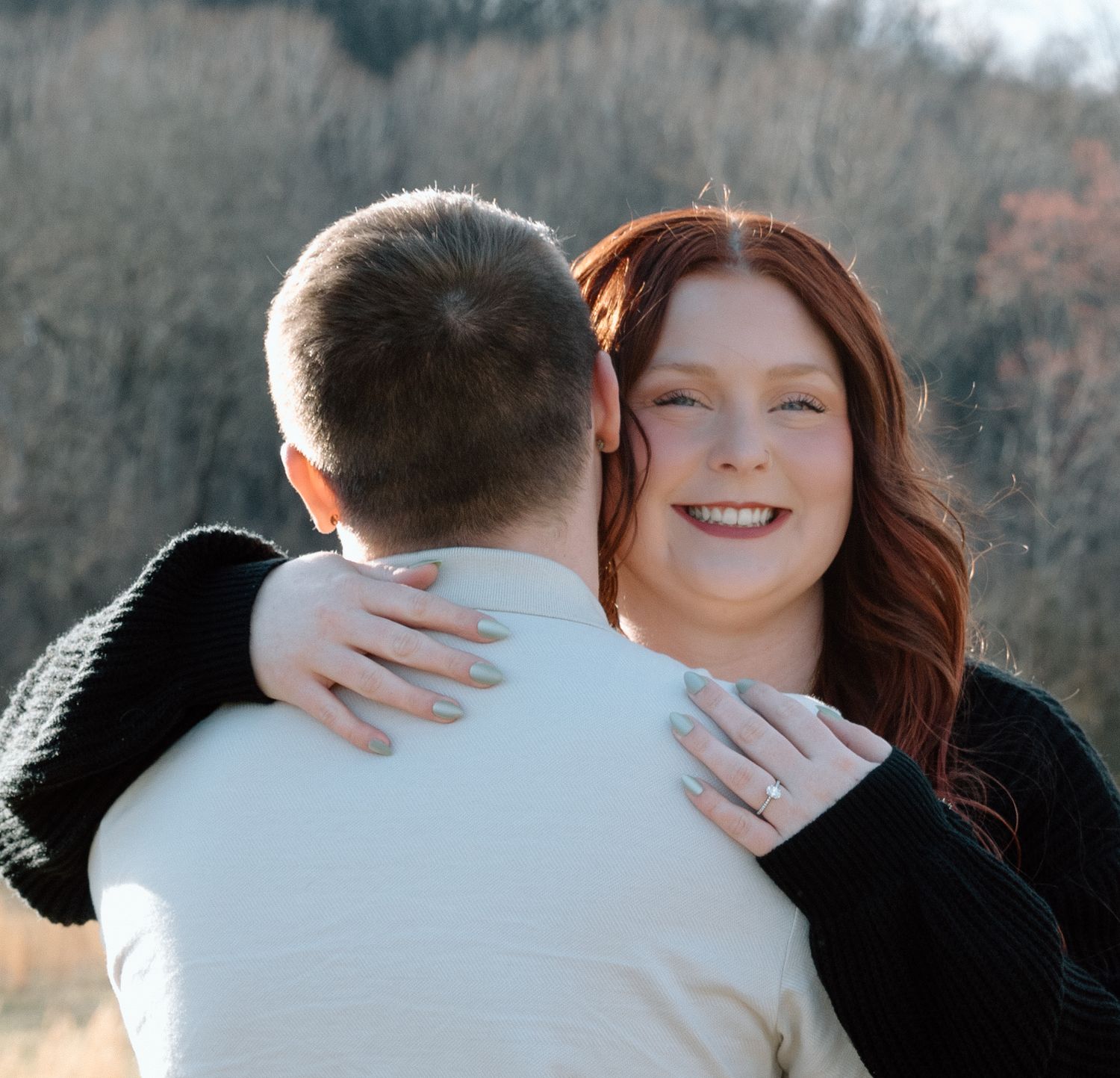 A smiling redhead woman hugs a man outdoors, showcasing an engagement ring, with bare winter trees in the background.