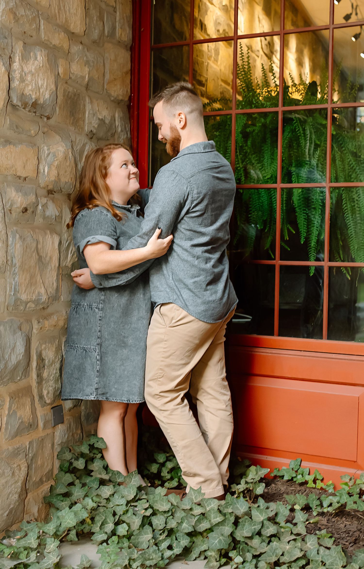A couple shares a romantic moment by a red-framed window on a stone building, surrounded by ivy and ferns.