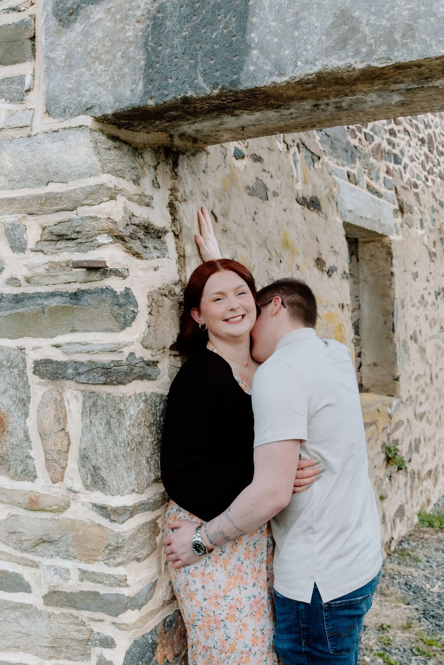 Couple embracing against a rustic stone wall, woman smiling joyfully as man kisses her cheek outdoors.