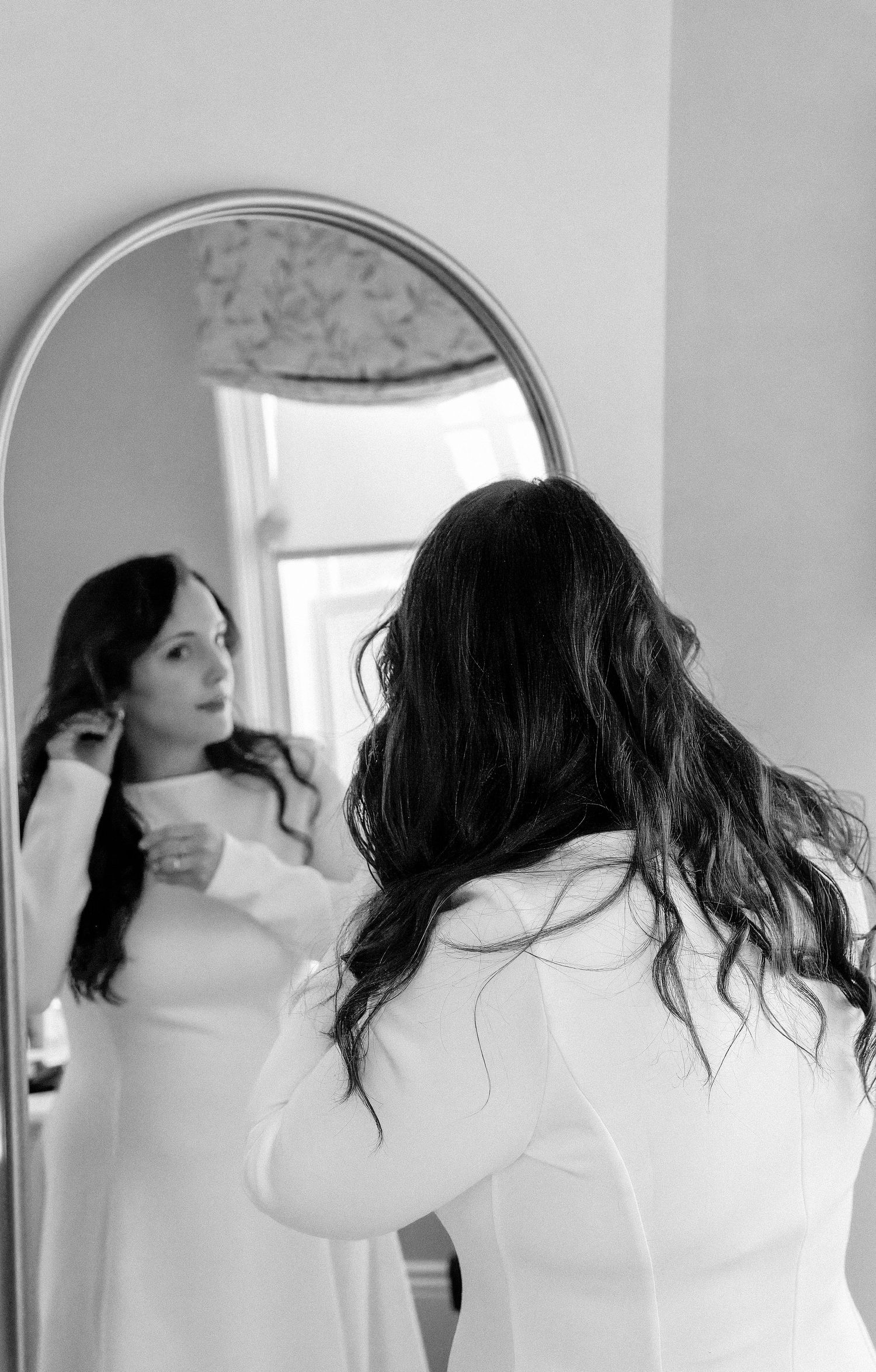 Black and white photo of a bride in a white dress adjusting her hair while looking into an arched mirror.