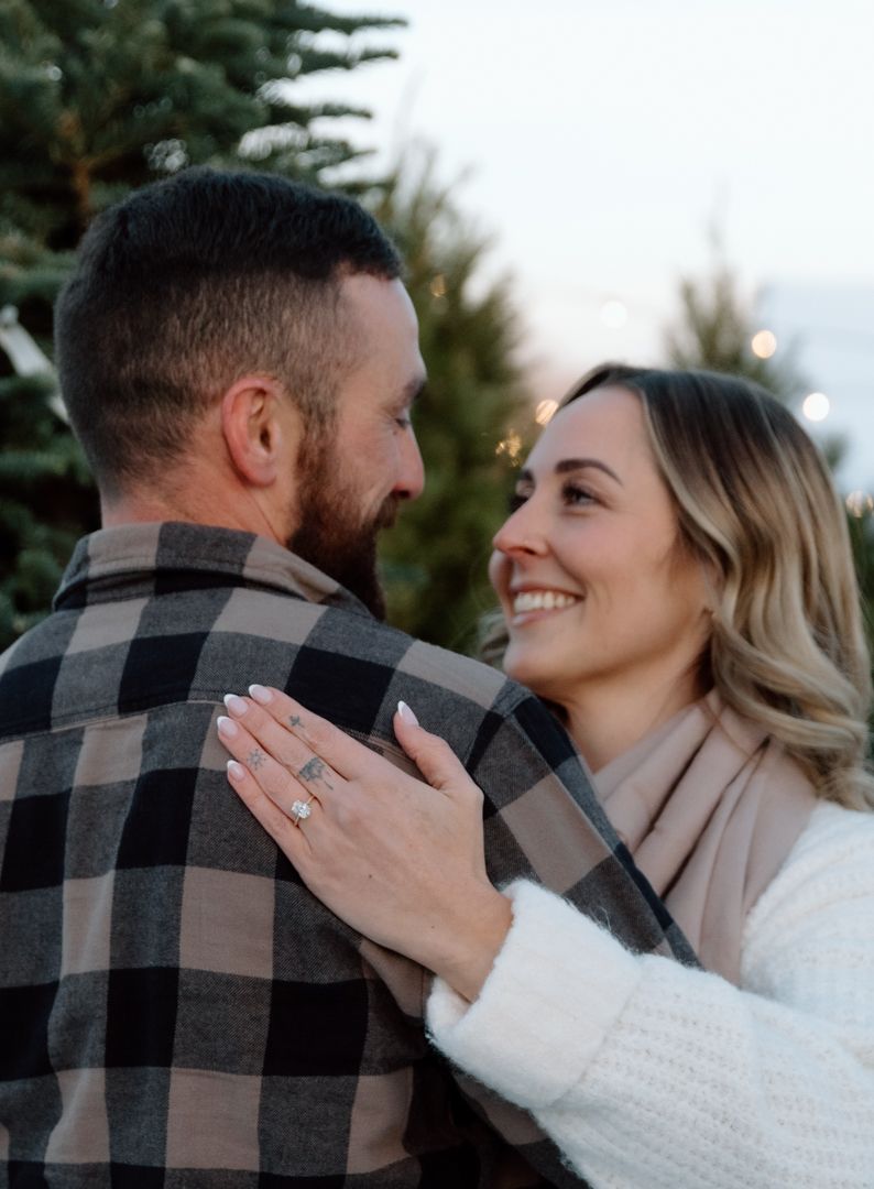 Couple embracing and smiling near Christmas trees, woman showing engagement ring on hand, wearing plaid and white sweater.
