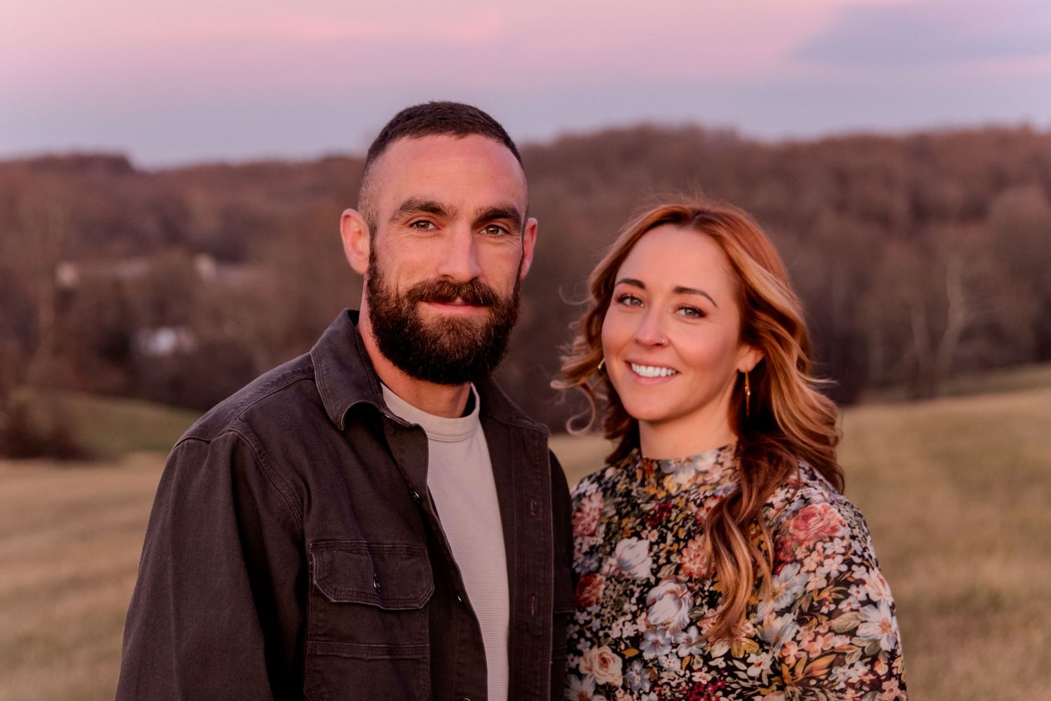 Couple smiling together outdoors at sunset with rolling green hills and a colorful sky in the background.