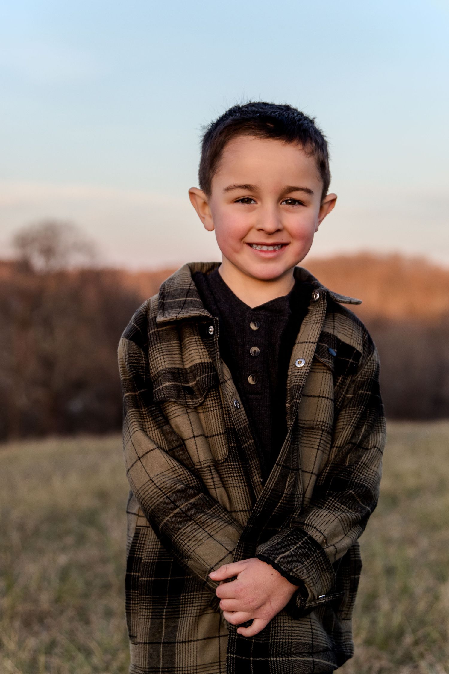 Child wearing plaid flannel jacket stands in outdoor field during golden hour with genuine smile.