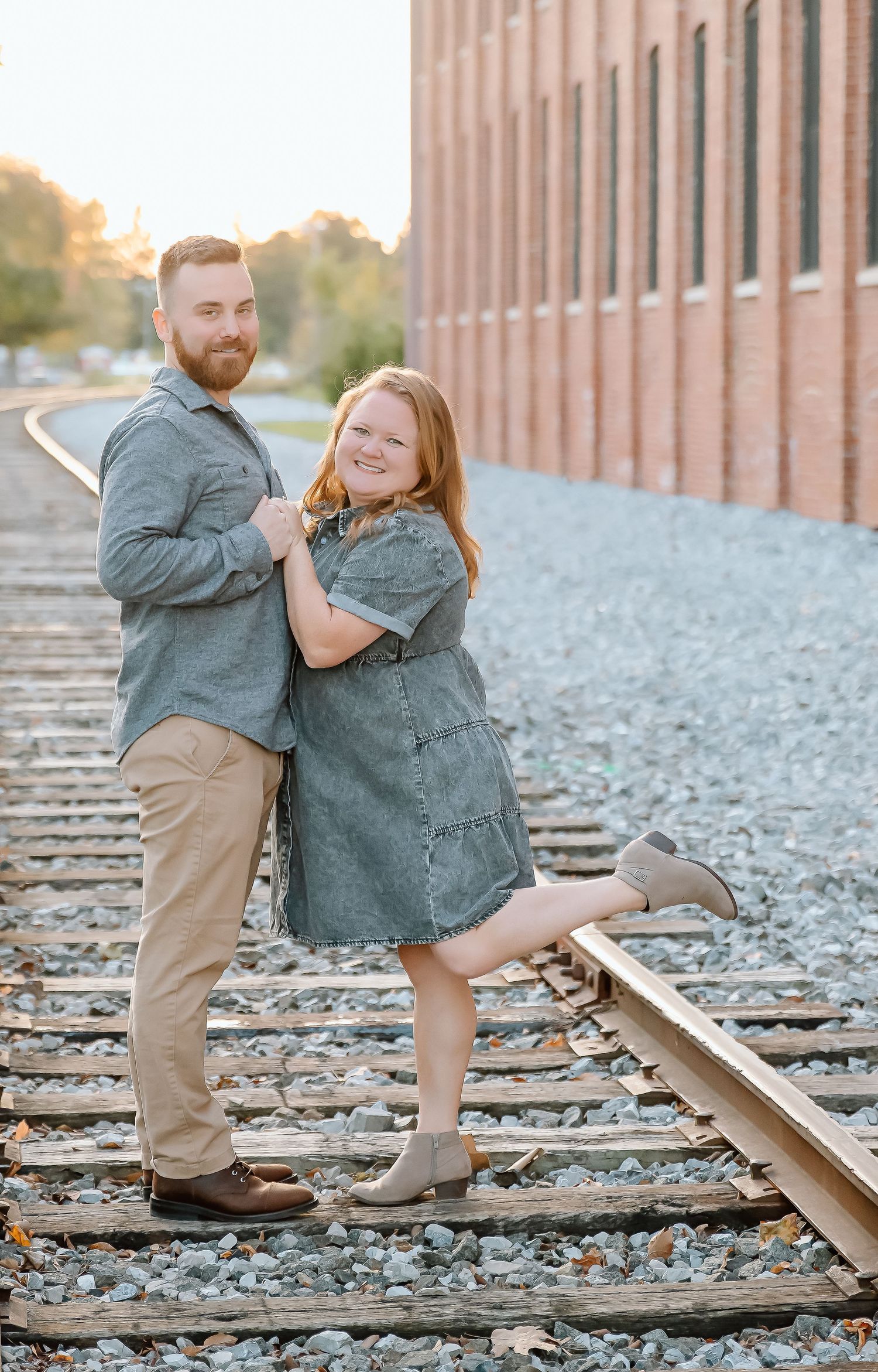 Couple posing on railroad tracks near brick building in matching gray outfits during golden hour photography session.