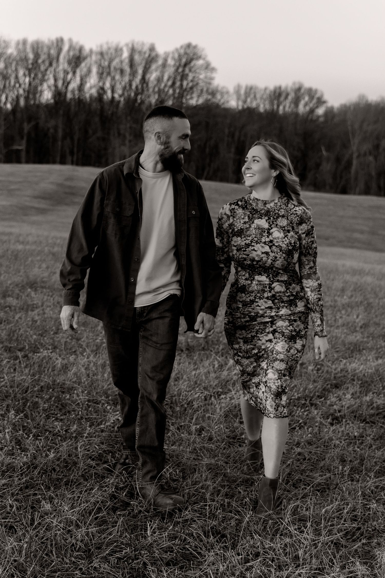 Black and white photo of a smiling couple holding hands and walking through a grassy field with trees in the background.