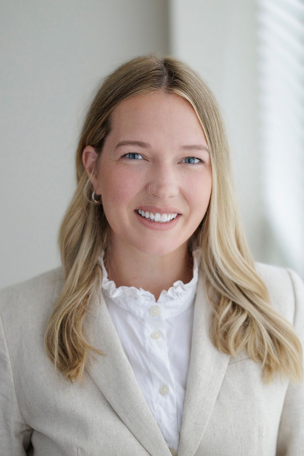 Smiling blonde woman in a beige blazer and white ruffled blouse, professional headshot with bright background.
