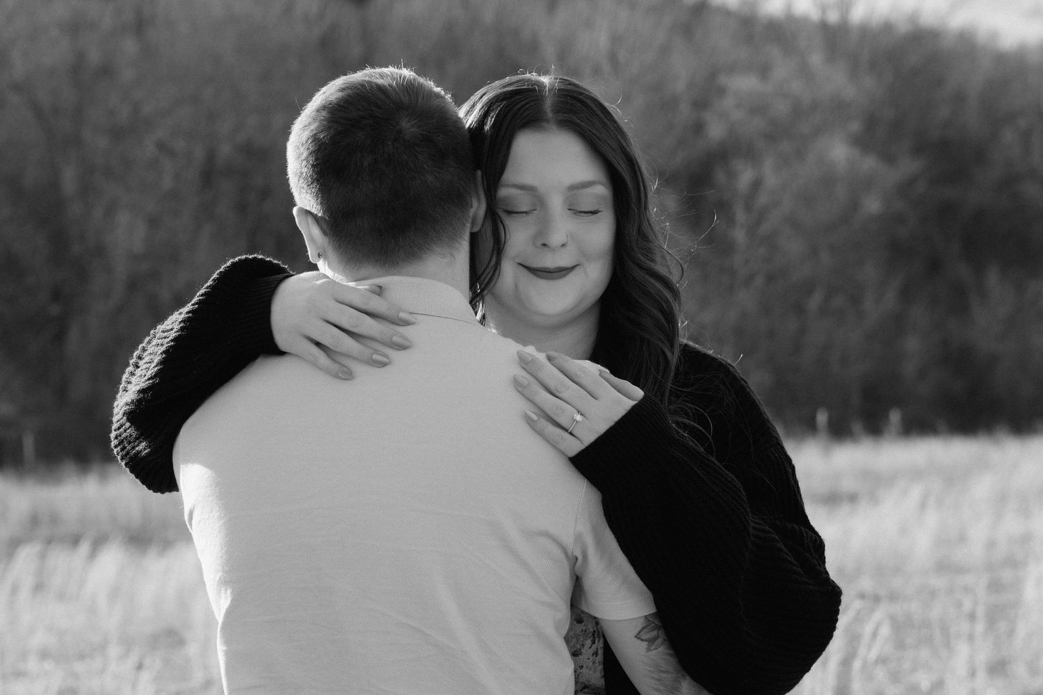 Black and white photo of a couple embracing outdoors, woman with eyes closed hugging man in a field.