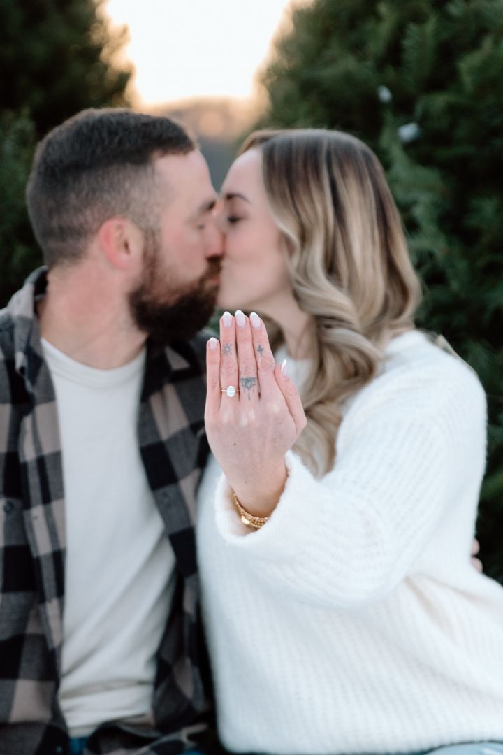 Couple kissing at Christmas tree farm, woman displaying diamond engagement ring on outstretched hand.