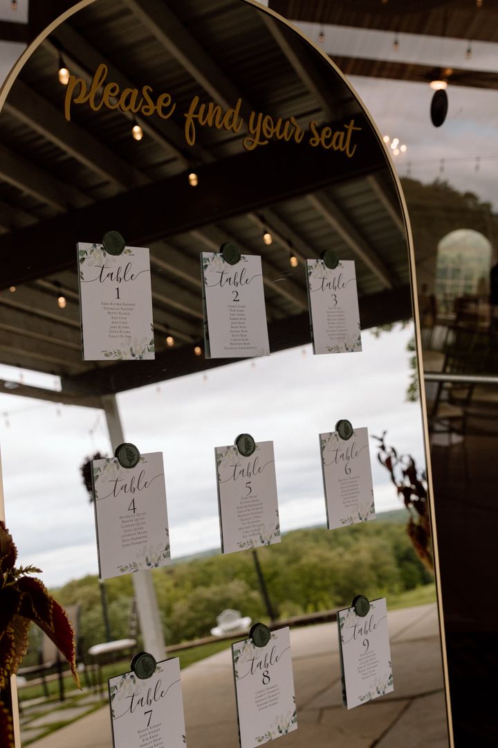 Wedding seating chart display with gold 'please find your seat' text and white paper cards hanging on a glass stand.