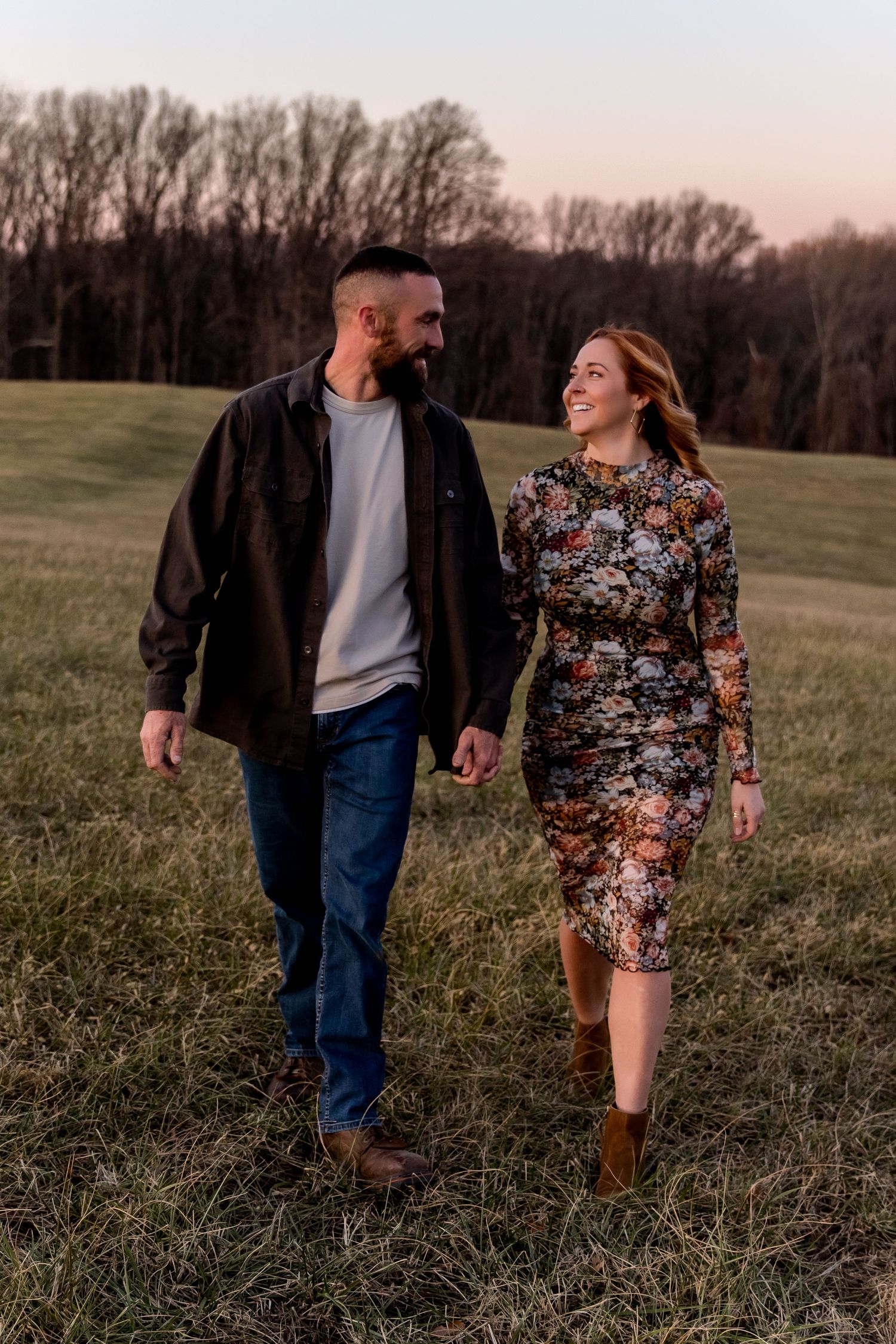 A couple walks together through a grassy field at sunset, with trees silhouetted in the background.