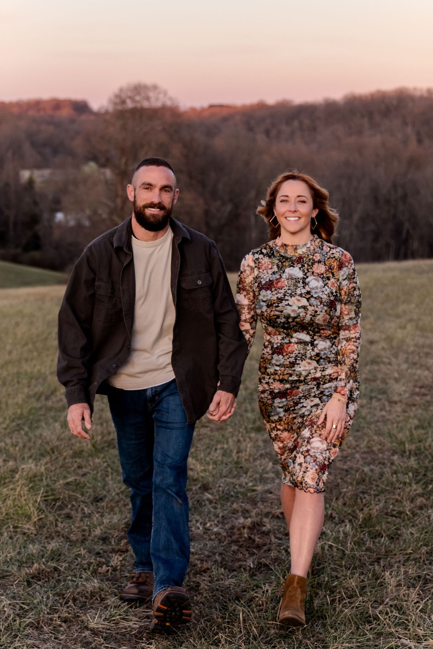 Couple holding hands walking in a field at sunset, man in dark jacket and jeans, woman in floral dress.