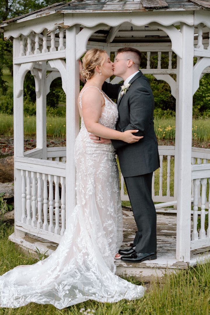 A romantic wedding couple shares a tender kiss under a white garden gazebo surrounded by lush green grass.