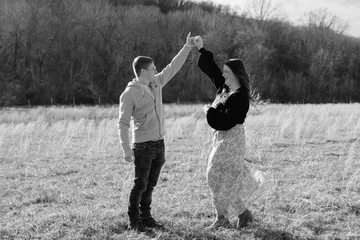 A couple dances together in a meadow on a sunny day with hills in the background.