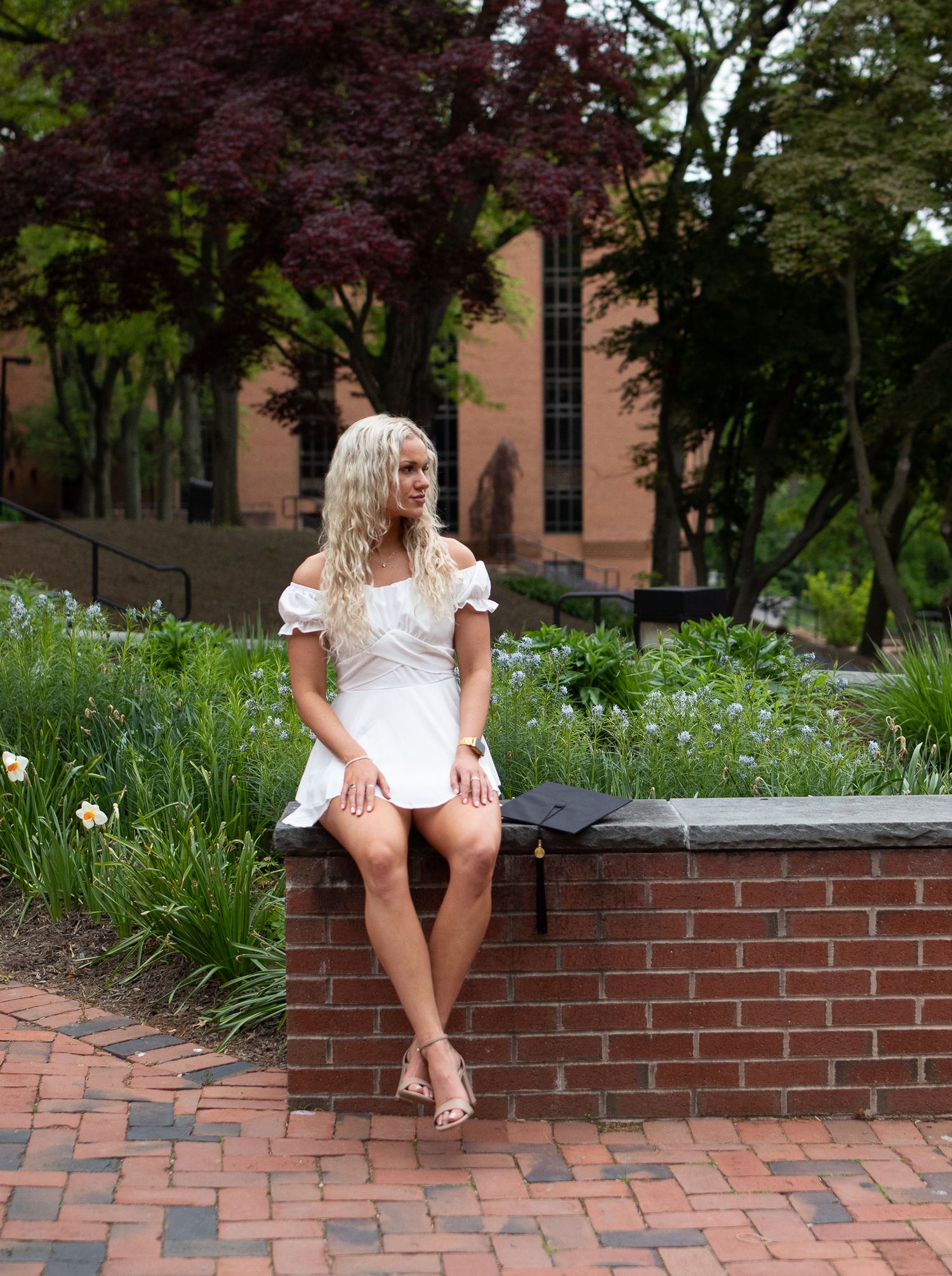 A figure in a white dress sits on a brick ledge surrounded by blooming flowers in a garden setting.