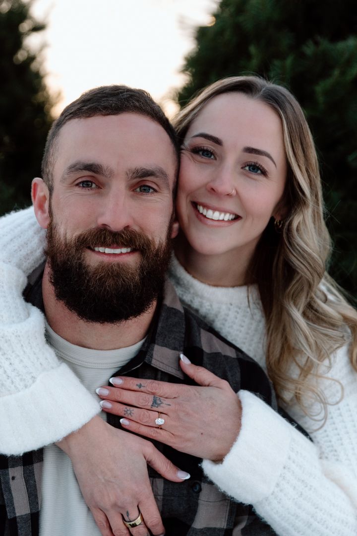 Smiling couple in white sweaters embrace outdoors near evergreen trees, woman showing engagement ring.