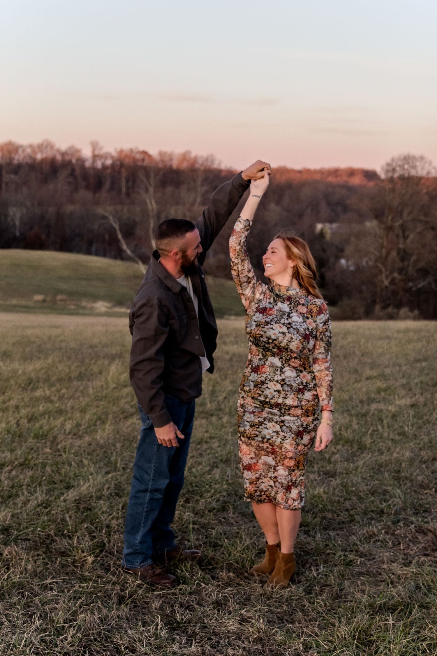 A couple shares a romantic moment in a grassy field at sunset while the person in a floral dress spins playfully.