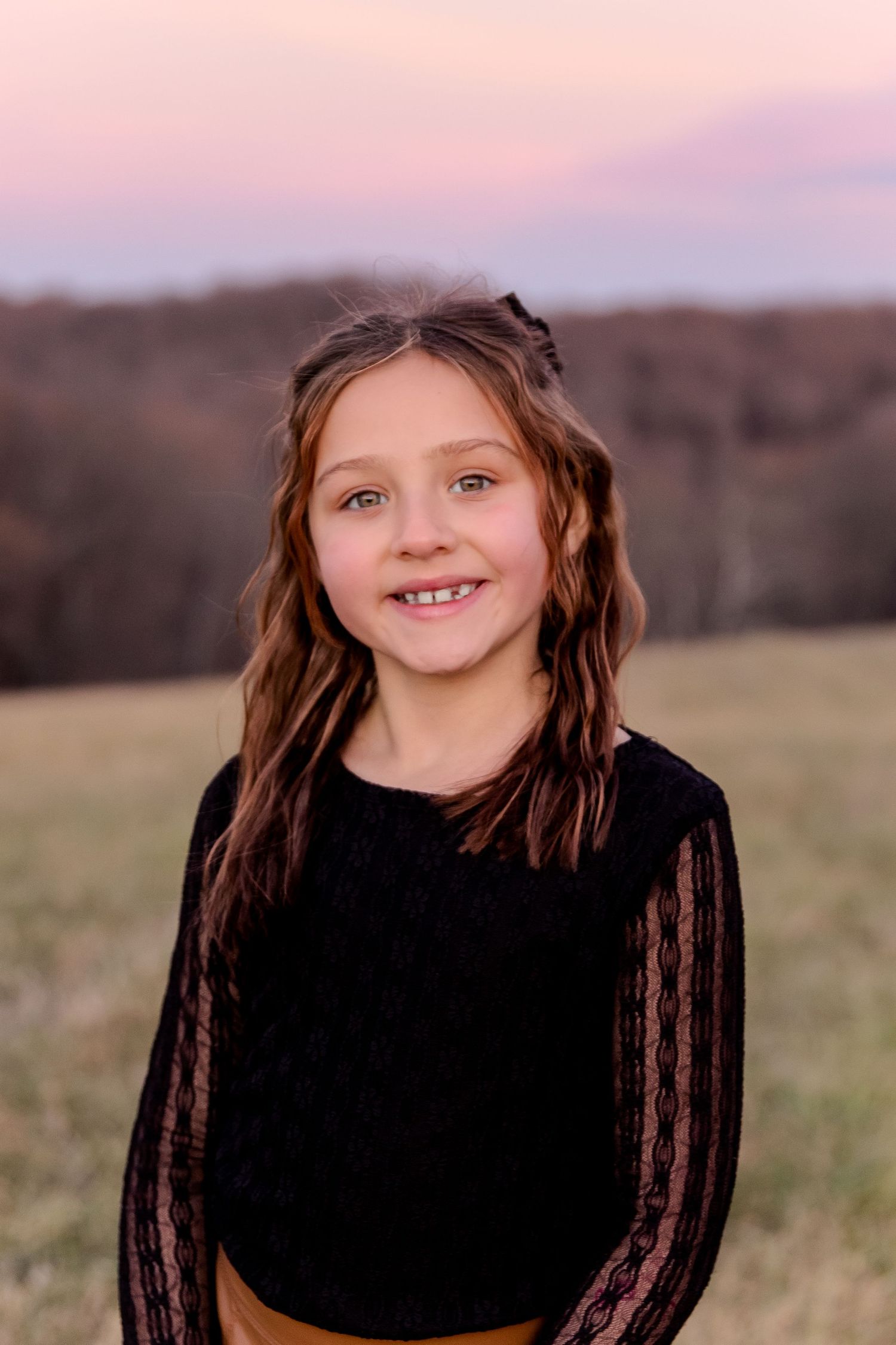 Young person in black sweater smiling at camera in outdoor natural setting with pink sunset sky.
