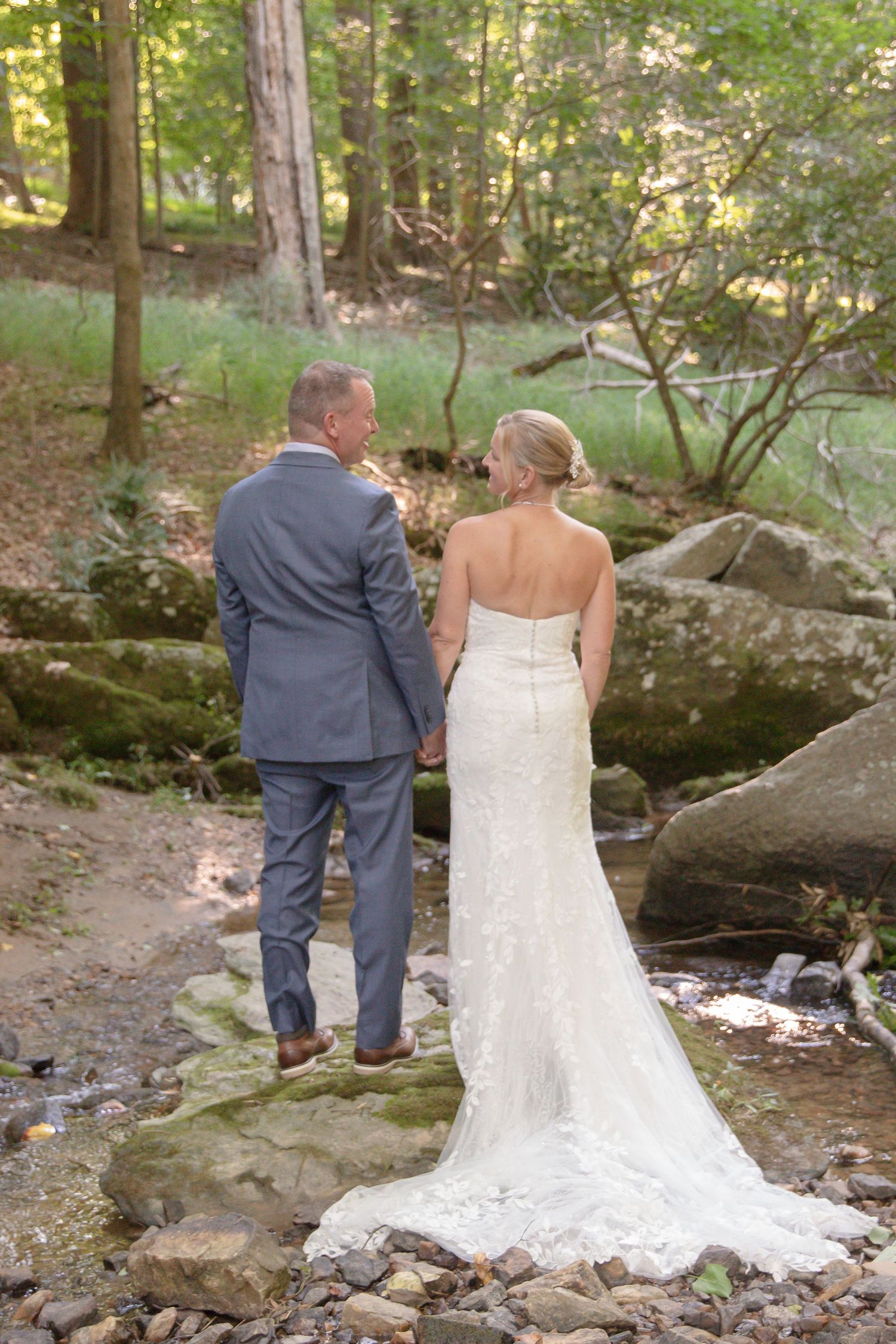 A couple in wedding attire sits together on a large boulder by a woodland stream in a serene forest setting.