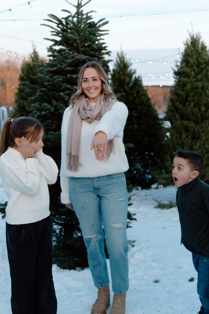 Woman in white sweater and scarf showing engagement ring to two surprised children at a snowy Christmas tree farm.