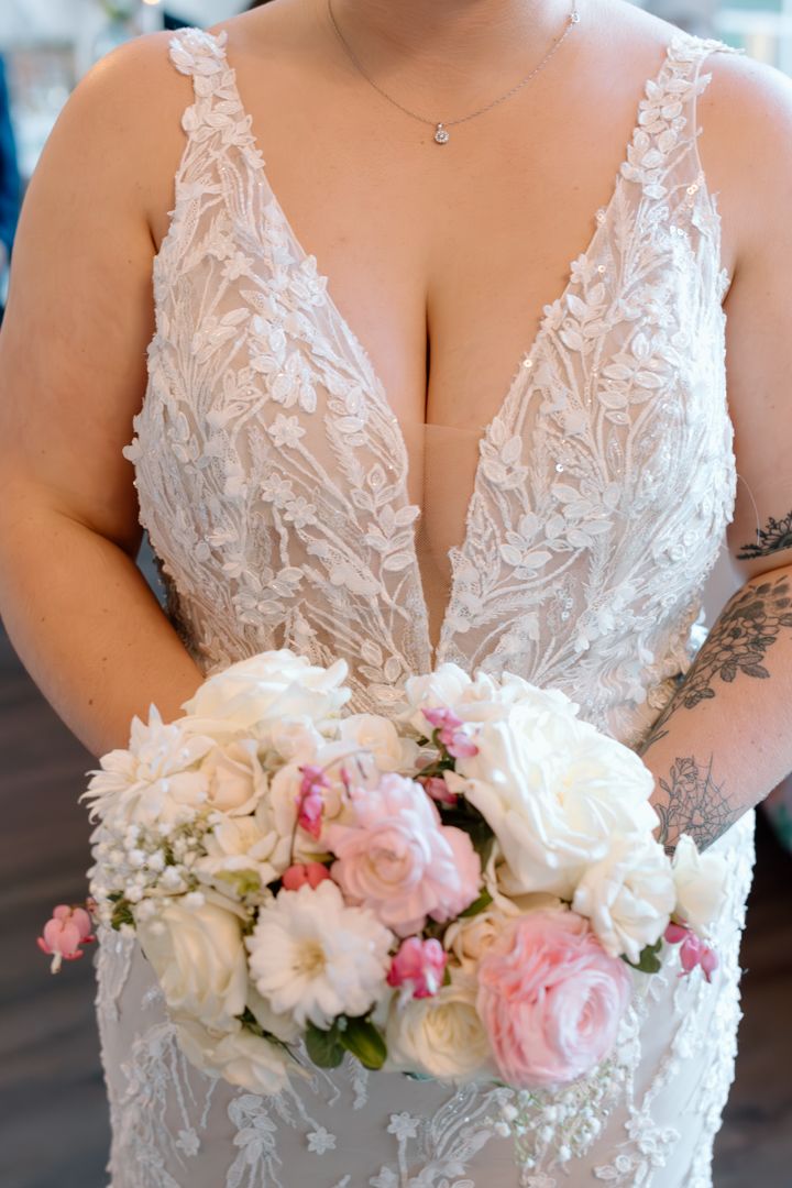 Close-up of white lace wedding dress bodice with delicate floral details and pink and white bouquet.