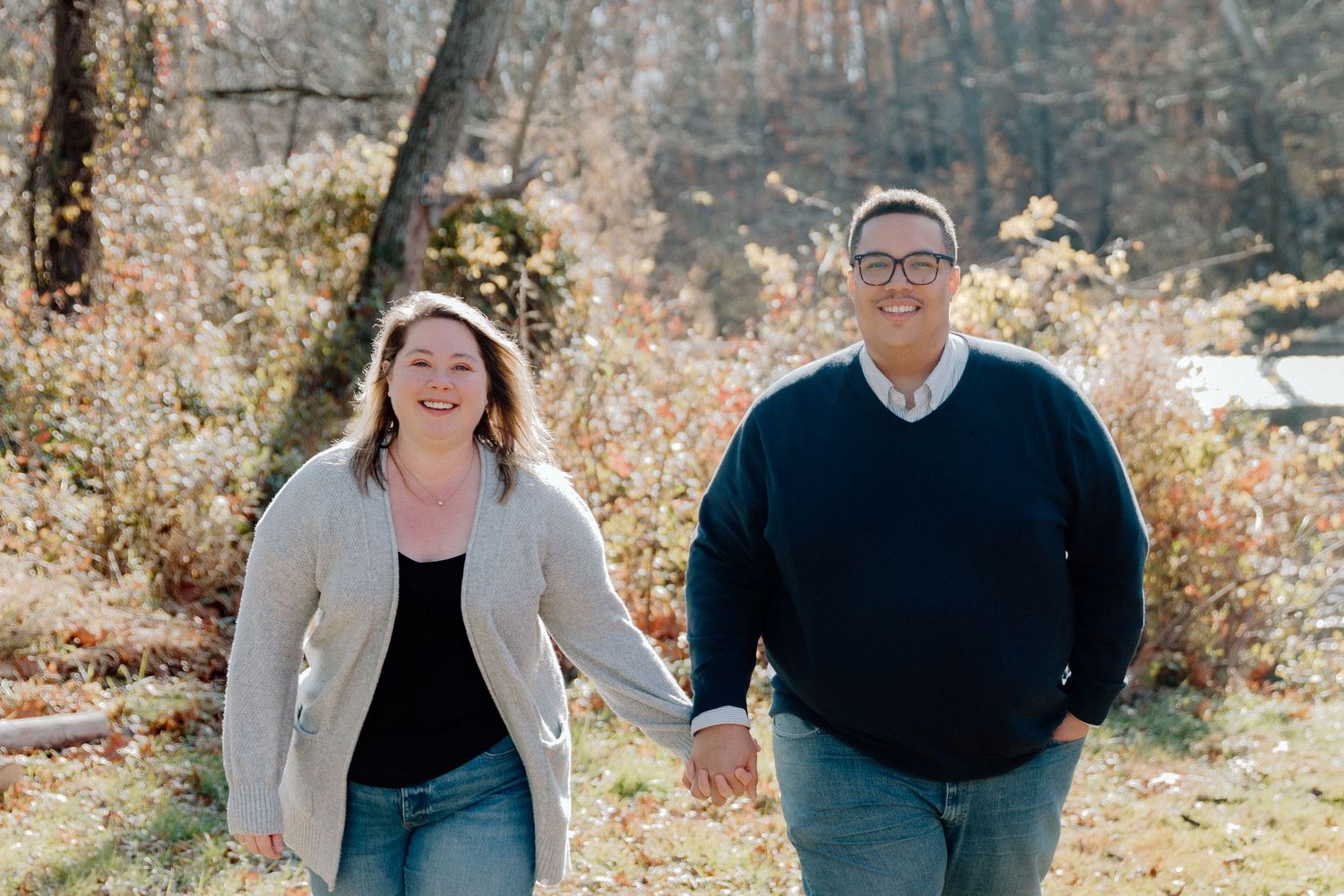 A happy couple walks along a scenic autumn path surrounded by golden foliage during an outdoor photo shoot.