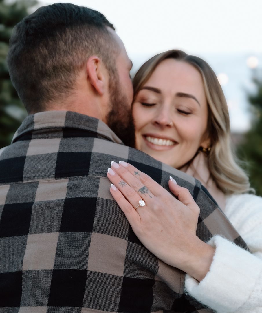 Smiling woman in white sweater hugs man in plaid shirt, showing off her diamond engagement ring.